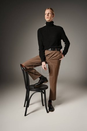 Foto de Plano vertical de atractivo joven no binario persona posando con pierna en silla negra, moda - Imagen libre de derechos