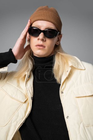 Vertikale Aufnahme eines jungen nicht binären Models mit Sonnenbrille und trendiger Winterjacke, das in die Kamera blickt