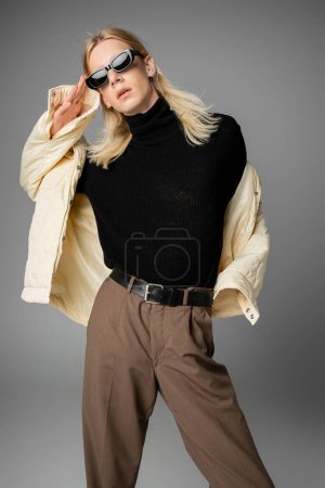 Foto de Joven modelo no binario de buen aspecto en chaqueta de invierno posando en gafas de sol y mirando a la cámara - Imagen libre de derechos