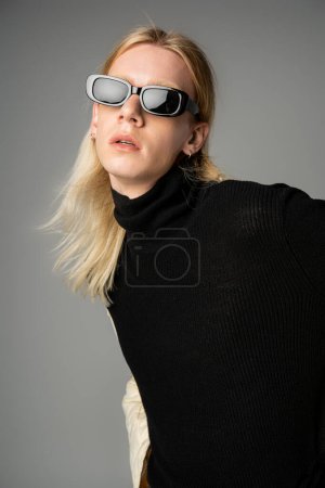 Foto de Joven persona no binaria en gafas de sol con estilo con el pelo largo posando sobre fondo gris, moda - Imagen libre de derechos