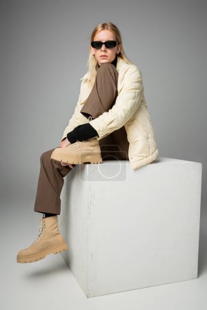 Photo for Young attractive non binary in stylish warm attire with sunglasses sitting on white huge cube - Royalty Free Image