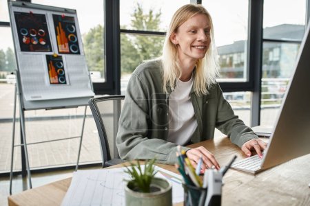 joyous non binary person working at table in office and smiling cheerfully, business concept