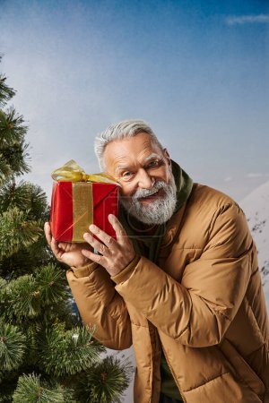 happy man dressed as Santa in warm jacket holding present near ear with mountains backdrop, winter