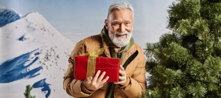 cheerful Santa holding present and smiling at camera with mountains backdrop, winter concept, banner