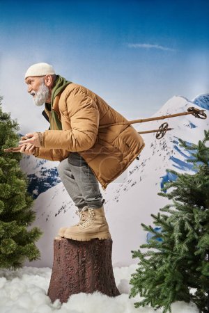 Sportlicher Weihnachtsmann hockt mit Skistöcken auf Baumstumpf vor Bergkulisse, Winterkonzept