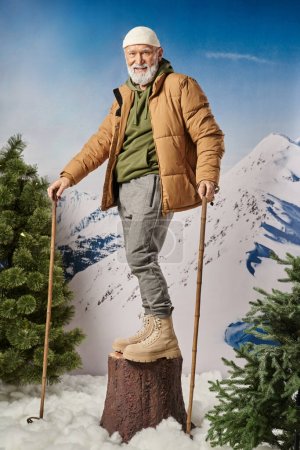Sportlicher Mann steht auf Baumstumpf und hält Skistöcke lächelnd in die Kamera, Winterkonzept