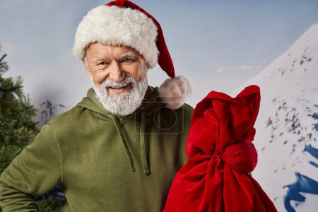 joyful athletic man in khaki hoodie wearing Santa hat and holding present bag, winter concept