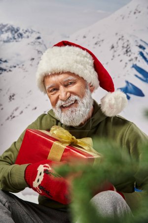 cheerful smiling Santa in christmassy hat and mittens holding present in hands, winter concept
