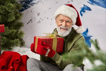 Sportlicher Weihnachtsmann mit rotem Hut und Fäustlingen sitzt mit Geschenk auf Schnee neben Geschenktüte, Winterkonzept