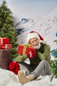 cheerful man dressed as Santa holding present and sitting on snow near tree stump, winter concept mug #681089014