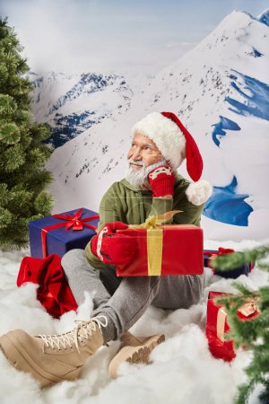 verträumter Mann im Weihnachtsmannkostüm auf Schnee sitzend, umgeben von Geschenken mit händennahem Gesicht, Winterkonzept