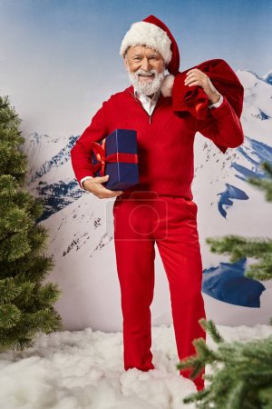 feliz Papá Noel moderno con barba blanca en traje rojo posando con regalo y bolsa de regalo, concepto de invierno