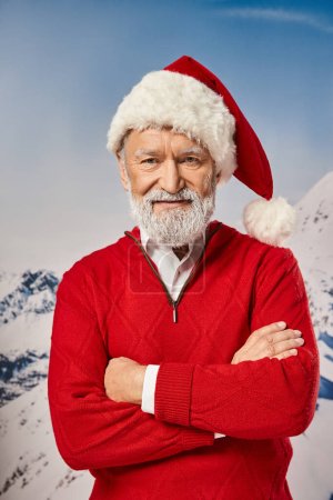 elegante Santa en traje rojo posando con los brazos cruzados en el pecho y mirando a la cámara, concepto de invierno
