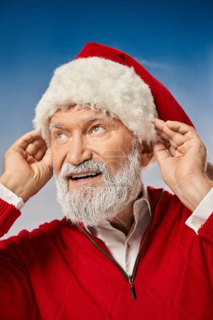 Porträt eines fröhlichen Mannes im Weihnachtsmannkostüm, der seinen weihnachtlichen Hut berührt und wegschaut, Winterkonzept