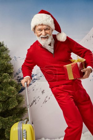 alegre traje de Santa con barba blanca posando con maleta amarilla y presente, concepto de Navidad