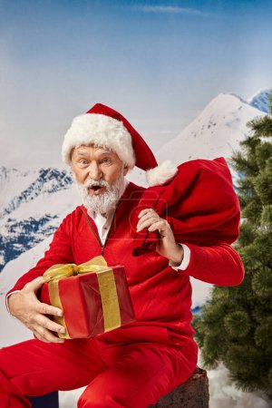 joyeux Père Noël avec sac cadeau et cadeau regardant la caméra avec bouche légèrement ouverte, concept d'hiver