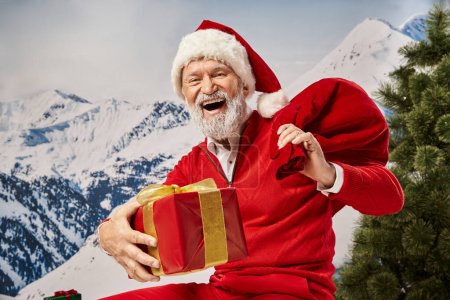 fröhlicher Mann im Weihnachtsmannkostüm, glücklich lächelnd in die Kamera, Geschenktüte und Geschenk haltend, winterliches Konzept