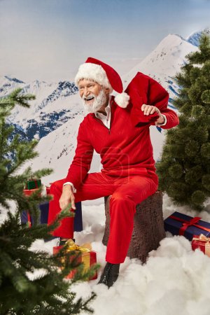glücklicher Weihnachtsmann sitzt auf Baumstumpf mit Geschenktüte neben Geschenken und schaut weg, Frohe Weihnachten