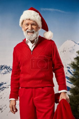 alegre Santa en traje rojo cálido posando con bolsa de regalo con telón de fondo de montaña, concepto de invierno