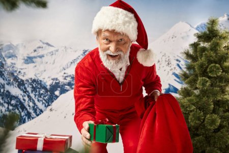 Père Noël ludique entouré de cadeaux montrant un à la caméra avec bouche légèrement ouverte, concept d'hiver