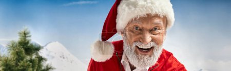 cheerful Santa in hat smiling happily at camera with mountain backdrop, winter concept, banner