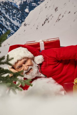 close up of joyous Santa in christmassy hat lying on snow surrounded by presents, winter concept