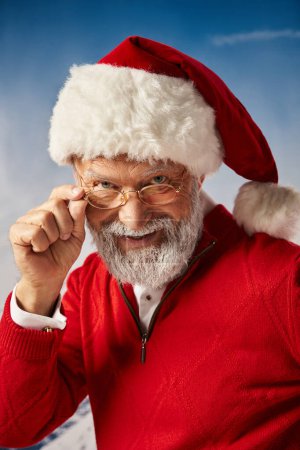 Foto de Feliz Navidad, alegre Santa en sombrero tocando sus gafas y sonriendo sinceramente a la cámara - Imagen libre de derechos