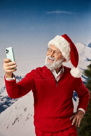 cheerful Santa with glasses taking selfie with one hand on hip with snowy backdrop, winter concept