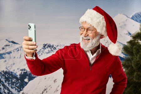 hombre feliz vestido como Santa con gafas y tomando selfie con telón de fondo de montaña, concepto de invierno