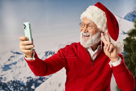 fröhlicher Weihnachtsmann mit weißem Bart in Brille macht Selfie und zeigt Friedensgeste, winterliches Konzept