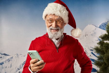 hombre alegre en traje rojo de Santa celebración de teléfono móvil y sonriendo a la cámara, concepto de invierno