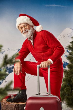 joyful man dressed as Santa in glasses posing with suitcase looking at camera, winter concept