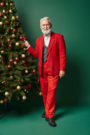 stylish handsome Santa Claus posing near Christmas tree touching decorations, winter concept