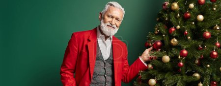 Photo for Elegant man dressed as Santa posing next to fir tree on green backdrop, winter concept, banner - Royalty Free Image