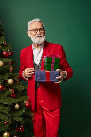 stilvoller Weihnachtsmann im schicken Anzug mit Geschenken und Blick in die Kamera vor grünem Hintergrund, Winterkonzept