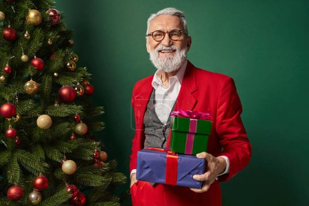 Téléchargez les photos : Homme élégant gai habillé en Père Noël avec des lunettes tenant des cadeaux à côté de sapin, concept d'hiver - en image libre de droit
