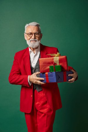 handsome man dressed as elegant Santa posing with presents on dark green backdrop, winter concept