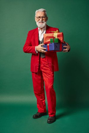 fröhlicher Weihnachtsmann im stilvollen roten Anzug mit Brille posiert mit Stapel von Geschenken, Winterkonzept