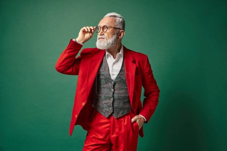 élégant Père Noël en costume rouge posant toucher ses lunettes d'une main dans la poche, concept de Noël