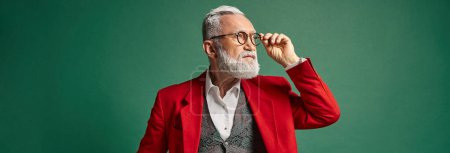 handsome Santa touching his glasses and looking away on green backdrop, winter concept, banner