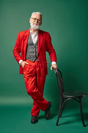 elegant stylish Santa with beard and glasses standing near chair and hand in pocket, winter concept