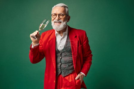 fröhlicher Mann als Weihnachtsmann verkleidet, der mit einer Hand in der Tasche ein Glas Champagner genießt, winterliches Konzept