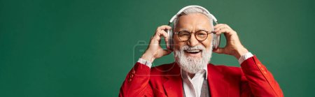 Téléchargez les photos : Homme élégant gai habillé en Père Noël posant avec des écouteurs sur fond vert, hiver, bannière - en image libre de droit