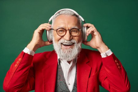 joyful stylish Santa in red elegant suit with headphones smiling at camera, winter concept