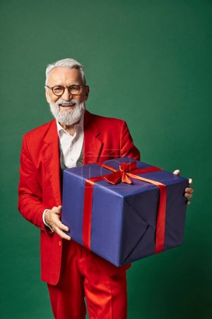 joyeux Père Noël en tenue rouge chic tenant un énorme cadeau et souriant à la caméra, concept d'hiver