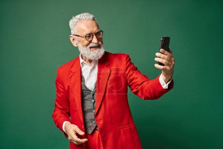 elegant good looking Santa in classy red attire taking selfie on green backdrop, winter concept