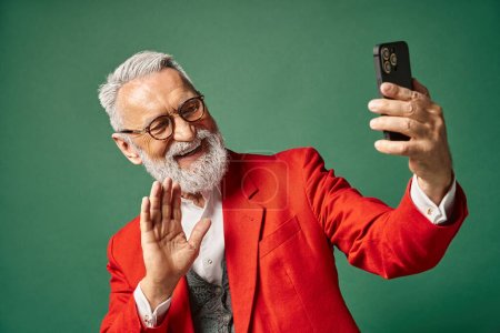 stylish jolly Santa with beard and glasses gesturing and waving at phone camera, winter concept