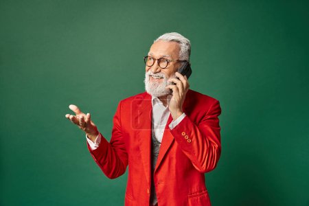 handsome Santa in stylish red attire gesturing and talking by phone cheerfully, winter concept