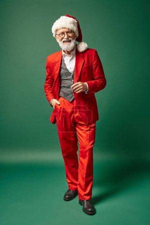 fröhlicher Mann als Weihnachtsmann gekleidet mit weihnachtlichem Hut posiert mit einer Hand in der Tasche, winterliches Konzept