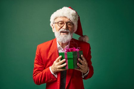 glücklich stilvoller Mann als Weihnachtsmann mit rotem Hut gekleidet hält Geschenk Blick in die Kamera, Winterkonzept